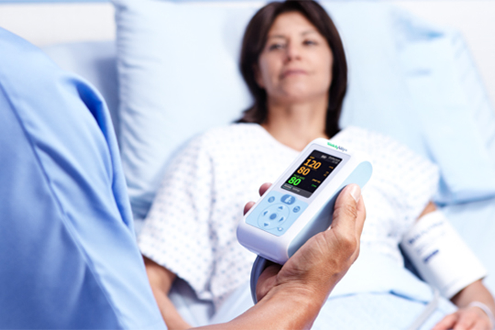 A clinician holds a Welch Allyn Connex ProBP 3400 Digital Blood Pressure Device. A patient lies in a hospital bed in the background.