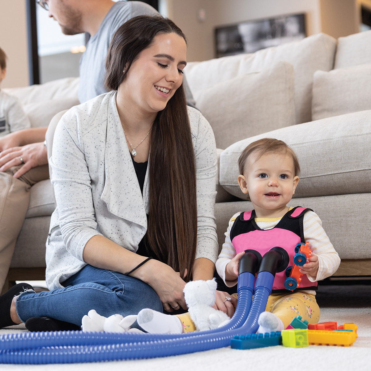 : Image of a woman sitting on the floor with her toddler, who is wearing a pink vest garment with hoses attached during treatment.